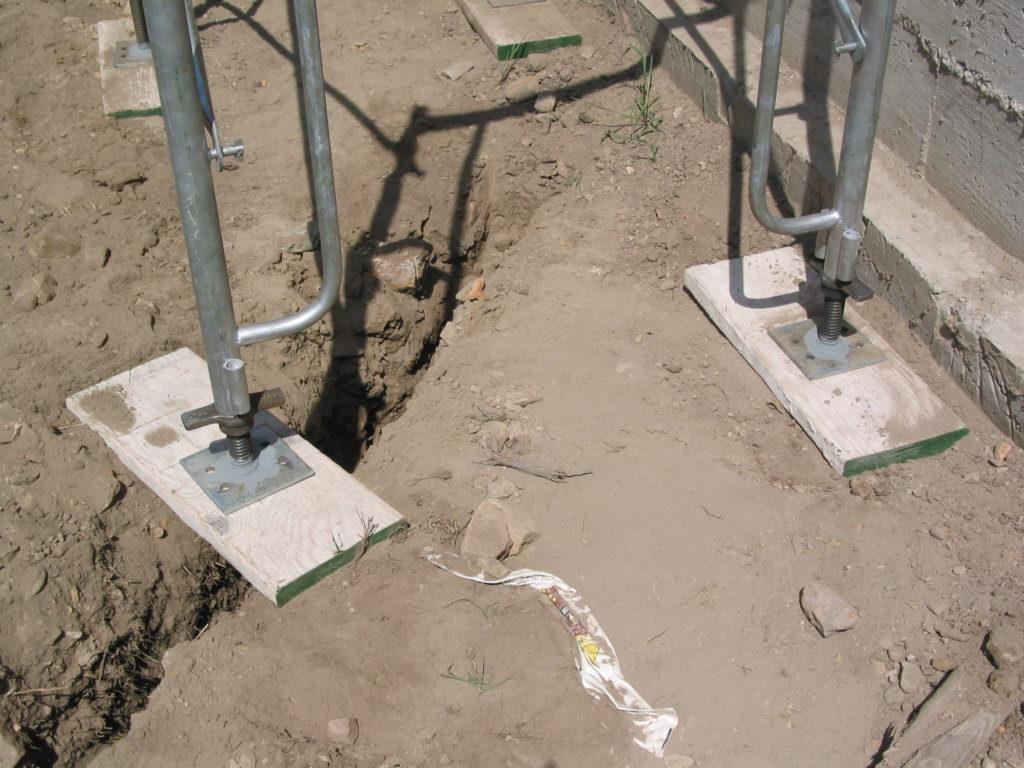 Scaffold screw jacks and base plates seated on timber mudsills over uneven, eroded soil beside a foundation wall